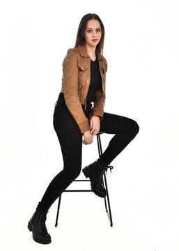 full length portrait of pretty brunette female model wearing basic modern  black clothing with brown leather jacket. sitting pose on chair, isolated figure on white studio background.
