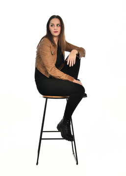 full length portrait of pretty brunette female model wearing basic modern  black clothing with brown leather jacket. sitting pose on chair, isolated figure on white studio background.
