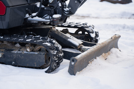Tracked mini excavator chassis on snow with dozer blade and hydraulic parts, close up of rubber track system, idler wheels and metal plow for cold season worksite operation