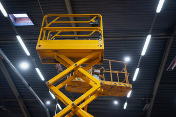 Yellow aerial work platform scissor lift extended high indoors, low angle shot with metal roof beams and bright linear lights, access equipment for construction maintenance © K