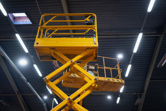 Yellow aerial work platform scissor lift extended high indoors, low angle shot with metal roof beams and bright linear lights, access equipment for construction maintenance