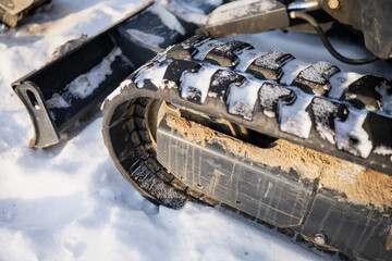 Winter close up of tracked construction machine showing rubber crawler track with snow buildup, dirt stains and undercarriage panel, industrial equipment detail in bright sunlight © K