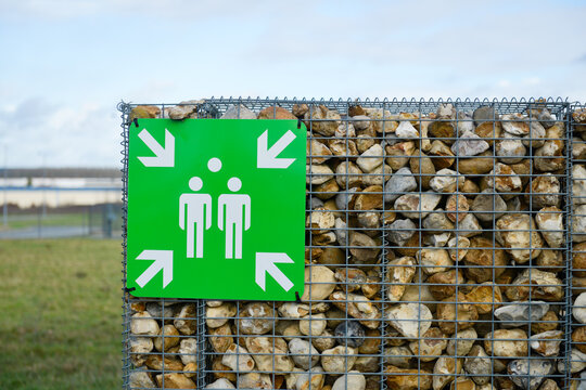 Panneau carr&eacute; sur fond vert illustrant un point de rassemblement avec quatre fl&egrave;ches pointant deux silhouettes, accroch&eacute; sur un mur de s&eacute;paration, compos&eacute; de roches dans un grillage