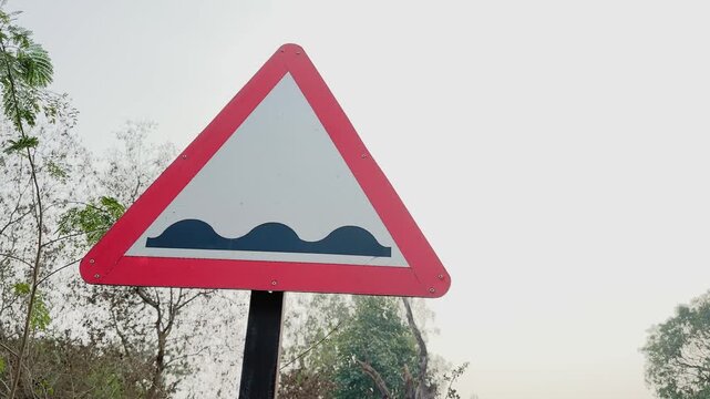 Camera orbiting around a triangular road humps ahead warning sign on a roadside pole, red border and wave symbol visible against pale sky and trees in daylight.