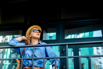 Naklejka premium Woman in hat and sunglasses leaning on metal railing looking at modern urban architecture