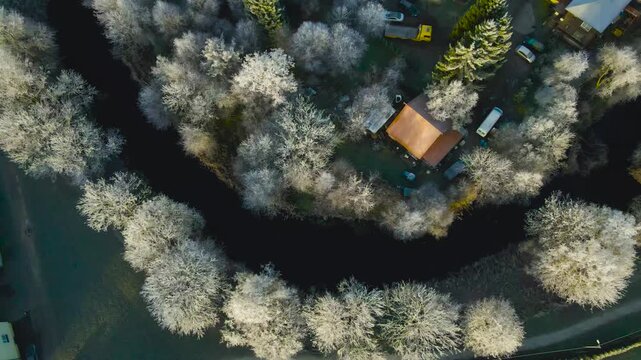 Top-down drone view of a dark river winding through frost-covered trees beside a red-roof cabin in golden winter light.