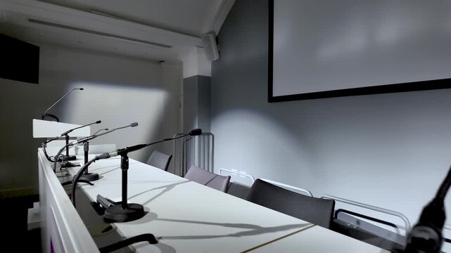 Empty Conference Hall With Microphones On White Table Presentation Screen And Chairs For Corporate Events Seminars And Public Speaking Engagements.