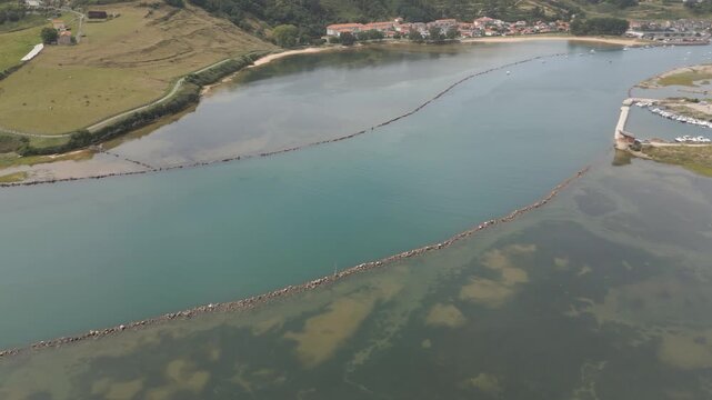 lateral drone flight focusing on the Suances estuary,bordered by stone riprap on both sides.The inner channel shows striking blue water,with a small dock and boats on one side and residential houses