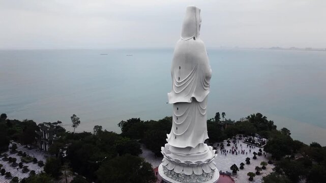 White majestic Lady Buddha statue, holding vase of holy water near coast, 360 drone shot.