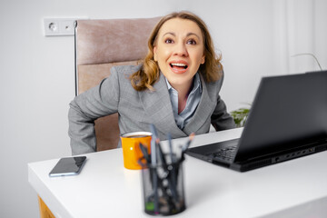 Excited young woman in grey blazer sitting at office desk with laptop looking up and laughing. Professional success, workplace happiness, career milestone, positive corporate culture.