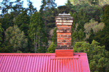 Traditional brick chimney on house with red corrugated metal roof with trees in background. Location: Auckland New Zealand