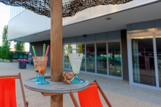 Bicchieri da cocktail e da gelato sono appoggiati su un tavolo di legno all'esterno di un bar. Il tavolo &egrave; fissato a un palo di legno e i bicchieri contengono cannucce colorate e dessert avanzati.