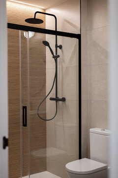 Modern bathroom with a glass shower enclosure, black fixtures, and beige tiles. A white toilet is visible in the corner.