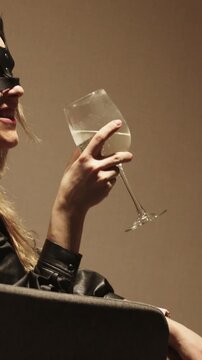 A woman in a black masquerade mask drinks alcohol from a glass and other woman playing with her with a feather tickler