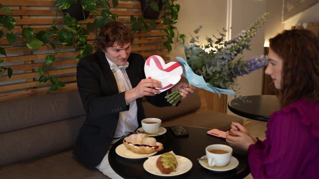 Shy man nervously presents flowers and heart shaped gift during romantic cafe date awkward body language quiet emotions and fragile connection on early stage relationship moment