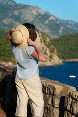 Young woman from behind adjusting her sun hat while standing at a viewpoint overlooking the mediterranean sea and picturesque mountain range.