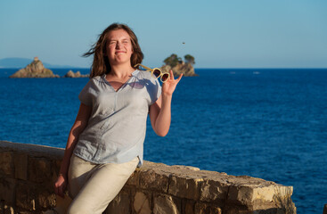 Joyful girl posing at the coast with sunglasses, enjoying the summer sunset light and sea breeze on her vacation trip.