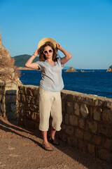 Young woman in straw hat and sunglasses posing on the viewpoint overlooking sea at sunset, summer resort vacation.