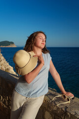 Young female traveler holding a sun hat and enjoying the evening glow at the mediterranean shore.