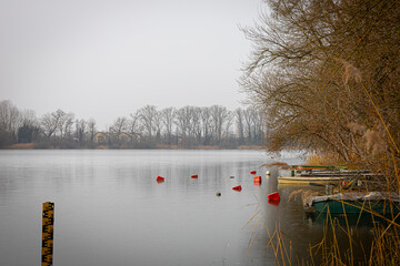 A serene winter landscape features a calm lake with scattered buoys and moored boats under an overcast sky. Bare trees line the banks, reflecting the quiet solitude of the season. © mirukser