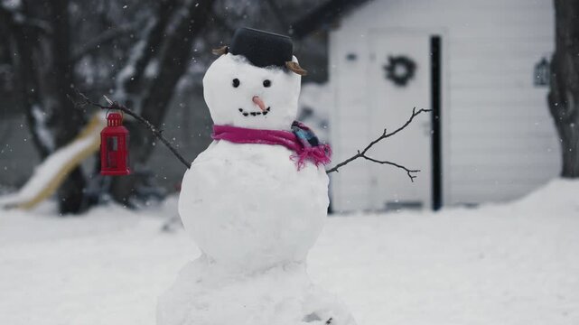 Snowman stands in yard with lantern during snowfall near house in winter season