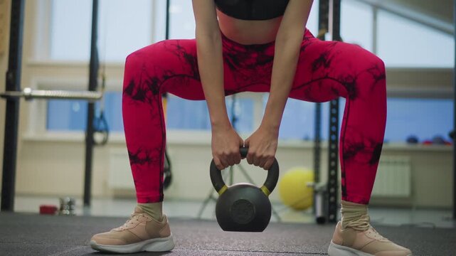 Female athlete in gym lifting kettlebell, wide sumo stance, bright leggings, focused lower body drive, controlled descent and powerful hip thrust, trainer demonstration vibe with studio equipment