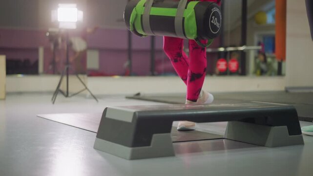 gym athlete performs weighted situps on mat with step platform and studio lights, vivid pink leggings and sandbag add intensity, coach and class in background, energetic pace and focused expression