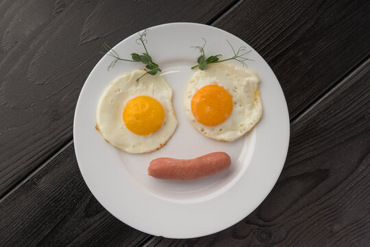 Two fried eggs and a sausage on a white plate, each egg garnished with a sprig of dill, dark surface.