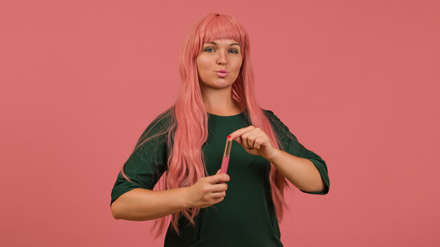 a woman in a pink wig looks at the camera making her lips into a bow while holding liquid lipstick