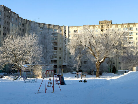 A winter city courtyard with gray multi-story panel buildings (Khrushchev-era or Brezhnev-era buildings) covered in a thin layer of snow and frost, under a bright blue sky