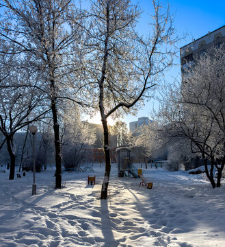 A winter city courtyard with gray multi-story panel buildings (Khrushchev-era or Brezhnev-era buildings) covered in a thin layer of snow and frost, under a bright blue sky