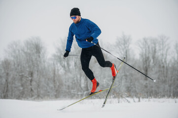 Male cross-country skier in blue jacket and black pants jumps over snow-covered terrain with trees in the background during winter sports activity