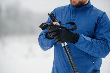 Male hiker in blue jacket adjusts trekking poles while standing in snowy landscape, blurred trees and soft snowfall visible in the background