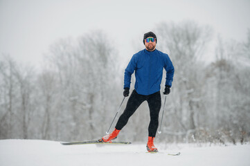 Male cross-country skier in blue jacket and black leggings glides on snowy trail surrounded by winter trees in a cold outdoor setting