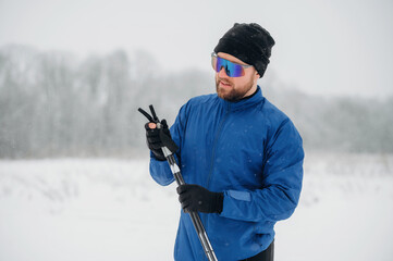 Male skier in blue jacket and black beanie adjusts ski poles while standing in snowy landscape with falling snowflakes and blurred trees in the background