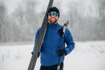 Naklejka premium Male skier wearing blue jacket and sunglasses holds ski equipment in snowy landscape with trees and falling snowflakes in the background