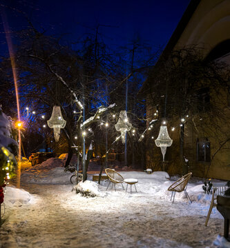 A summer courtyard, buried in snow on a winter evening: a cozy nook with swings, a slide, and benches, completely covered in fresh snow under the warm glow of streetlights. Gray multi-story buildings 