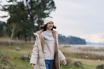 Obraz premium Woman outdoors in a winter coat walking in a park, wearing a sweater and beanie, enjoying a chilly day by the lake with calm landscape and serene nature