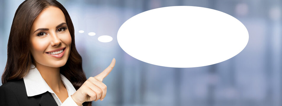 Happy smile cute business woman wear black suit jacket, white shirt, showing pointing with hand. Brunette businesswoman against blurred office background. Empty mockup speech bubble with free ad space