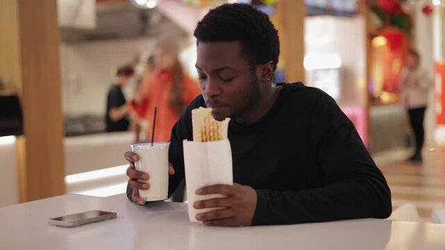 Young african male use phone and enjoying snack shaverma with chicken and milkshake at food court, fast food in mall shop.