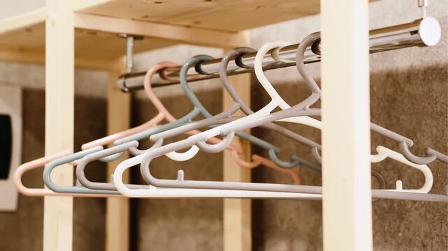 A close-up of a row of plastic coat hangers in pastel shades (beige, grey, white, peach) hanging neatly on a metal rod. Soft lighting creates a warm, minimalist atmosphere.