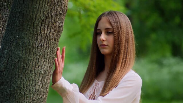 Standing next to a tree in slow motion, a girl softly moves her fingers very softly across the bark with a calm expression. Warm backlight and blurred greenery enhance the serene green forest setting.