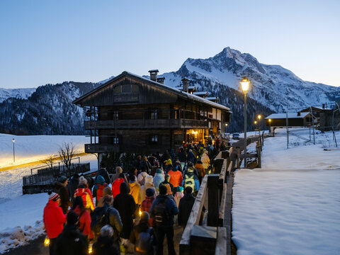 During the carnival procession, the people with lanterns walk across a bridge in a snowy landscape, with a large wooden building and mountains in the background. Sauris,Friuli Venezia Giulia,Italy
