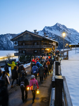 During the carnival procession, the people walk along a path at dusk, each carrying a lantern. A wooden building and snow-covered mountains are in the background. Sauris,Friuli Venezia Giulia,Italy