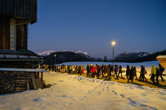 During the mountain carnival procession in Sauris, a group of people walk along a snowy path at dusk, illuminated by a street lamp, with mountains in the background. Sauris,Friuli Venezia Giulia,Italy