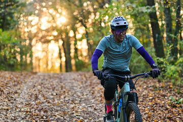 Obraz premium Mountain Biker Riding Through Autumn Forest Path With Helmet, Gear, and Sunlit Leaves