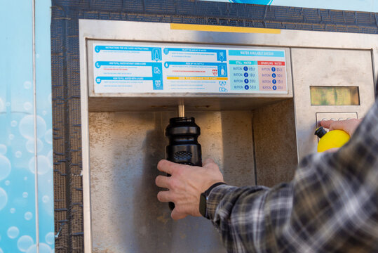 Person refilling a reusable bottle at an outdoor public water dispenser. Ideal for content on sustainability, eco-friendly urban living, and reducing single-use plastic waste.