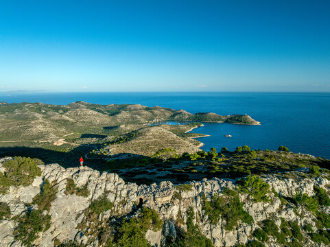 Aerial view of a lone figure standing atop a rugged, rocky cliff overlooking the stunning Adriatic Sea, where islands meet the clear blue horizon, Lastovo, Dubrovnik-Neretva County, Croatia.