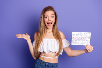 Excited young woman holding a marked calendar against a purple background while presenting an...