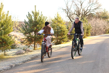 Fototapeta premium A teen and man ride bicycles side by side on a sun dappled rural road, frosty grass, evergreens, and warm light capture intergenerational bonding, shared joy, and peaceful motion.
